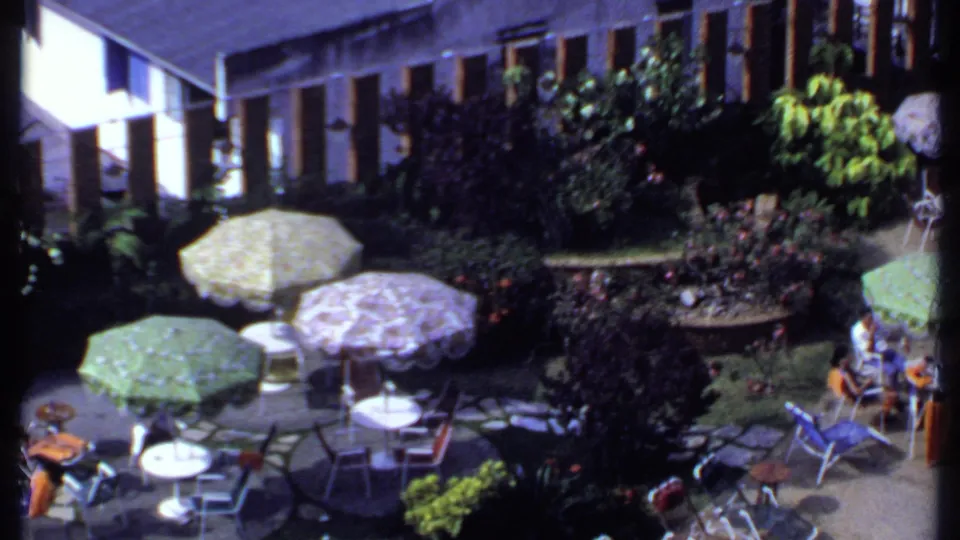 1969 India home-movie frame showing the patio gathering from above, with shaded seating, dense plants, patterned umbrellas, and guests near the garden edge.