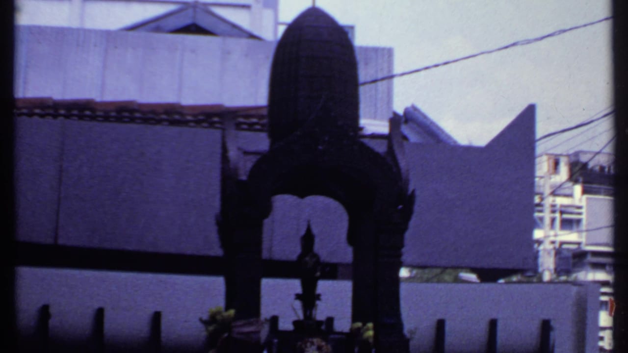1969 India home-movie frame showing a dark metal shrine from below, with concrete walls, roof tiles, wires, and building edges behind it.