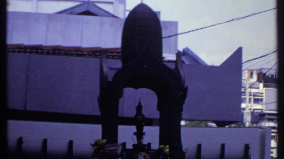 1969 India home-movie frame showing a dark metal shrine from below, with concrete walls, roof tiles, wires, and building edges behind it.