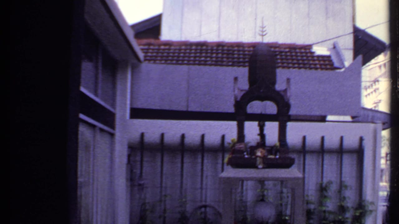 The final 1969 India frame widens to show the dark metal shrine on a concrete block, with an iron fence, pale walls, tiled roof, and building edges nearby.