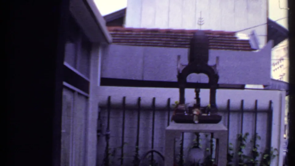 1969 India home-movie frame showing a dark shrine-like structure behind a fence in front of a house with a tiled roof.