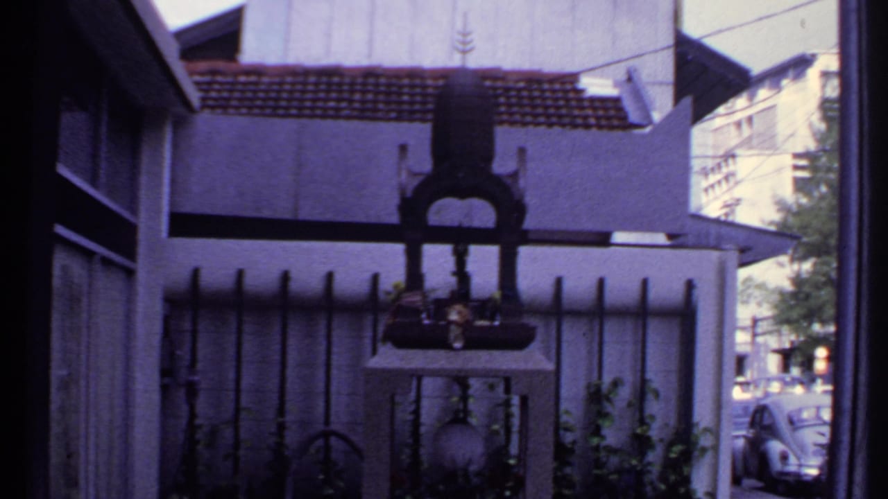 1969 India frame of a fenced shrine-like object outside a house, with plants and a parked car visible along the street.