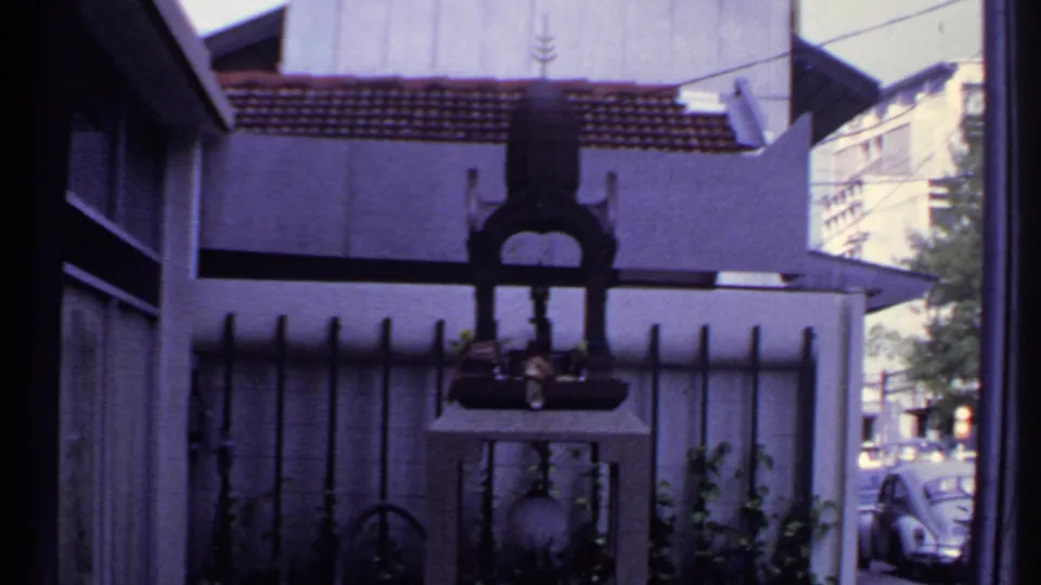 1969 India frame of a fenced shrine-like object outside a house, with plants and a parked car visible along the street.