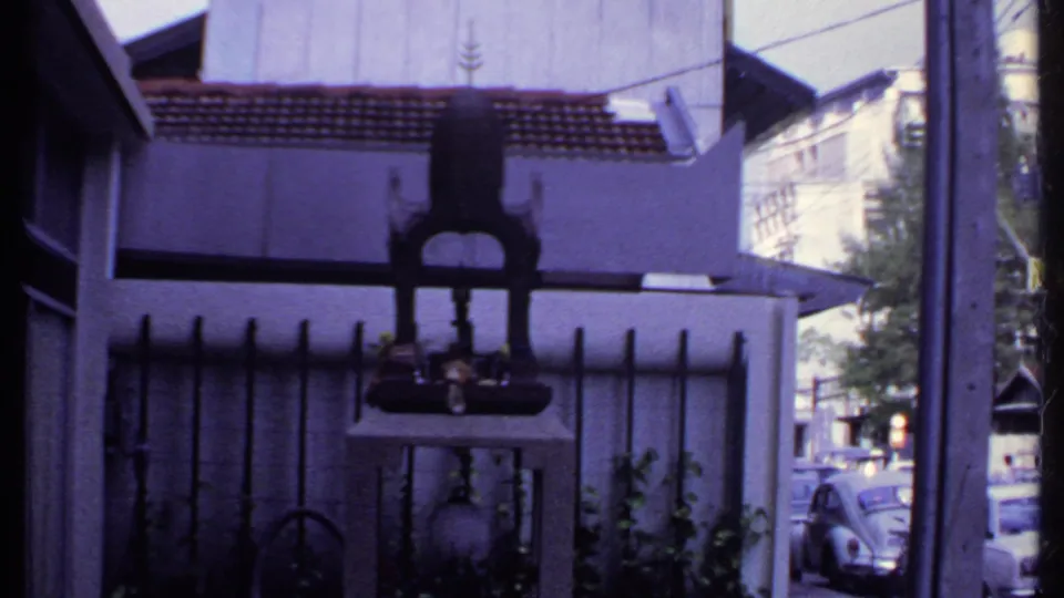 Final 1969 India frame showing the shrine-like object, house exterior, fence bars, plants, and parked cars beyond the street.