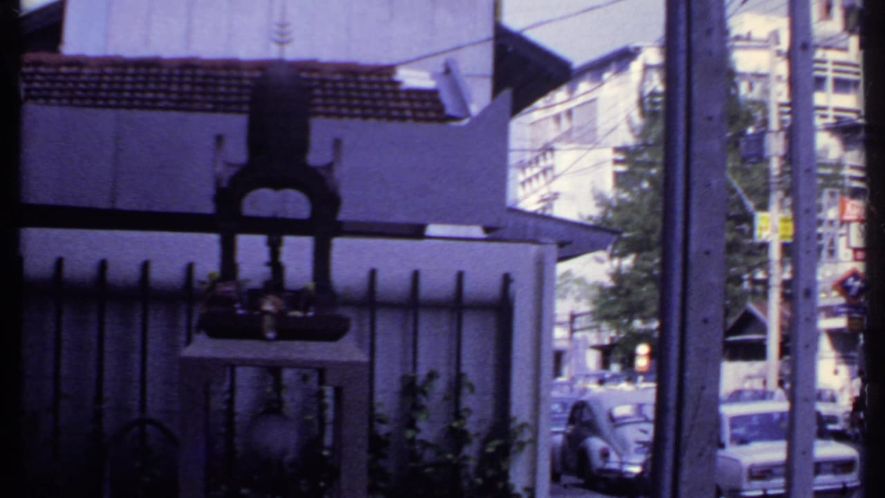 1969 India street view from behind a metal gate and shrine-like structure, with classic cars and storefronts beyond.