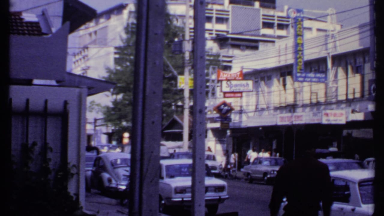 1969 India street view showing cars, pedestrians, shop signs, and a gated foreground as the camera pans across the block.
