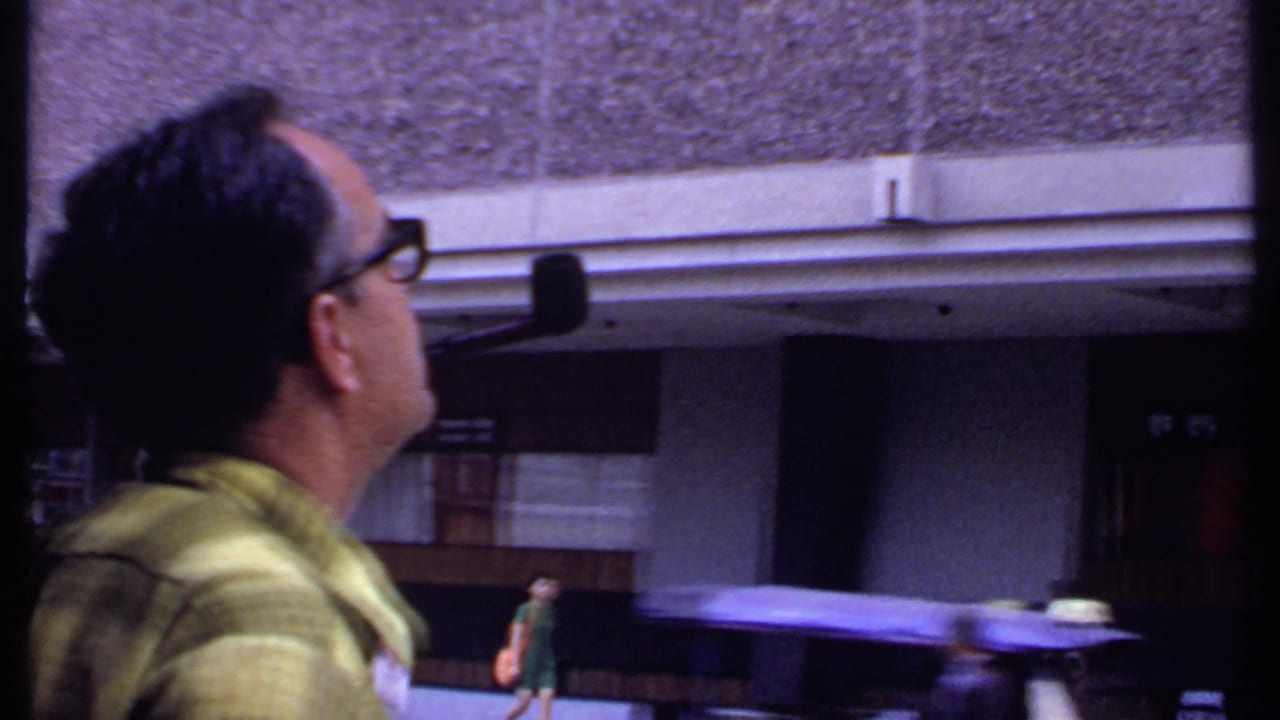 1969 India home-movie frame of a man in glasses and a green shirt looking upward near a street and building entrance.