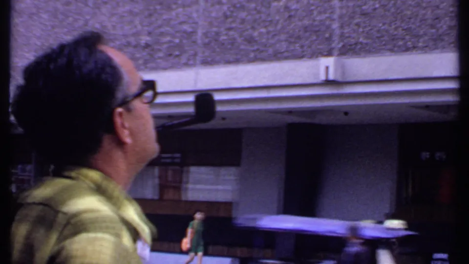1969 India home-movie frame of a man in glasses and a green shirt looking upward near a street and building entrance.