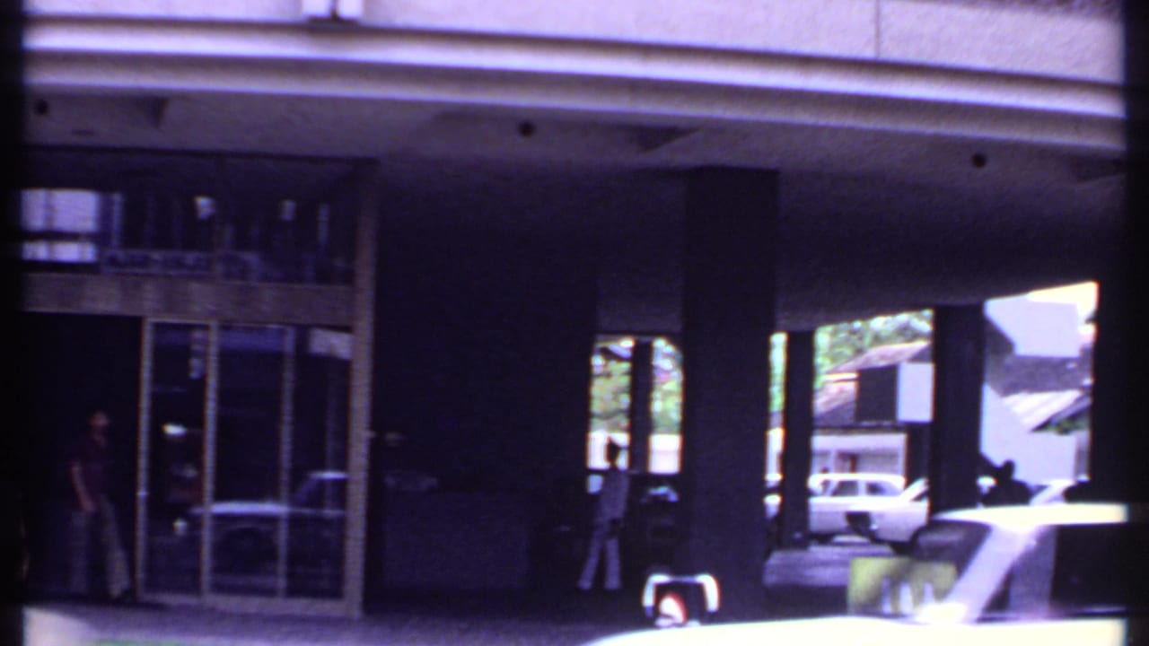 1969 India 8mm frame showing a modern covered entrance with pillars, glass doors, pedestrians in shadow, and a car moving past.