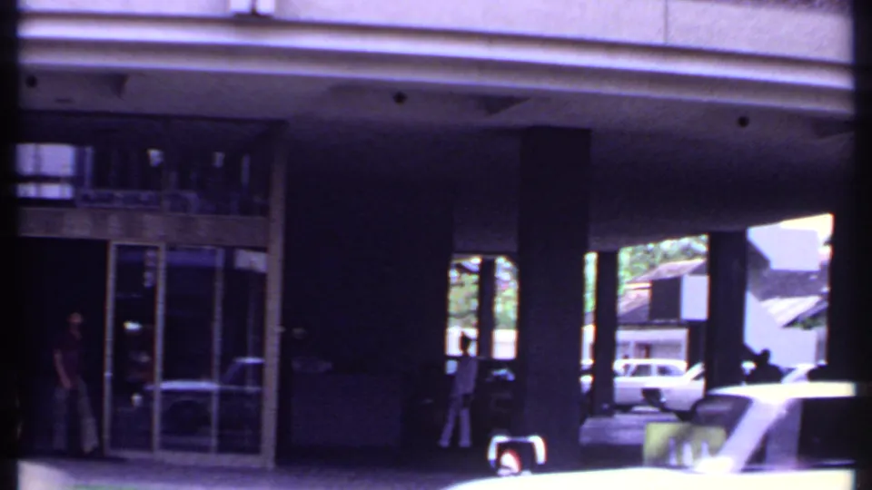 1969 India 8mm frame showing a modern covered entrance with pillars, glass doors, pedestrians in shadow, and a car moving past.