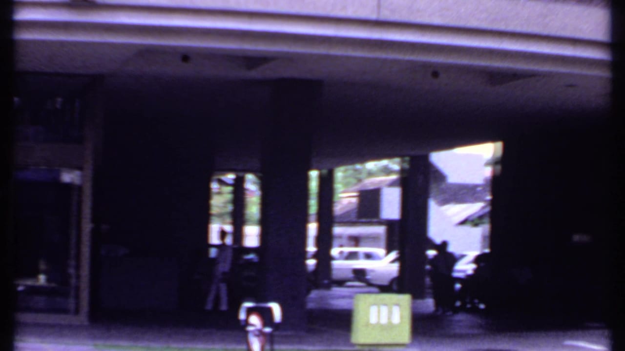 Building portico with pillars, a green roadside sign, pedestrians, and cars beyond the entrance.