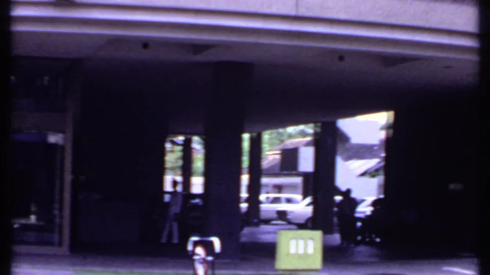 1969 India street view from 8mm film with a shaded building portico, green sign, pedestrians, and cars visible through the opening.