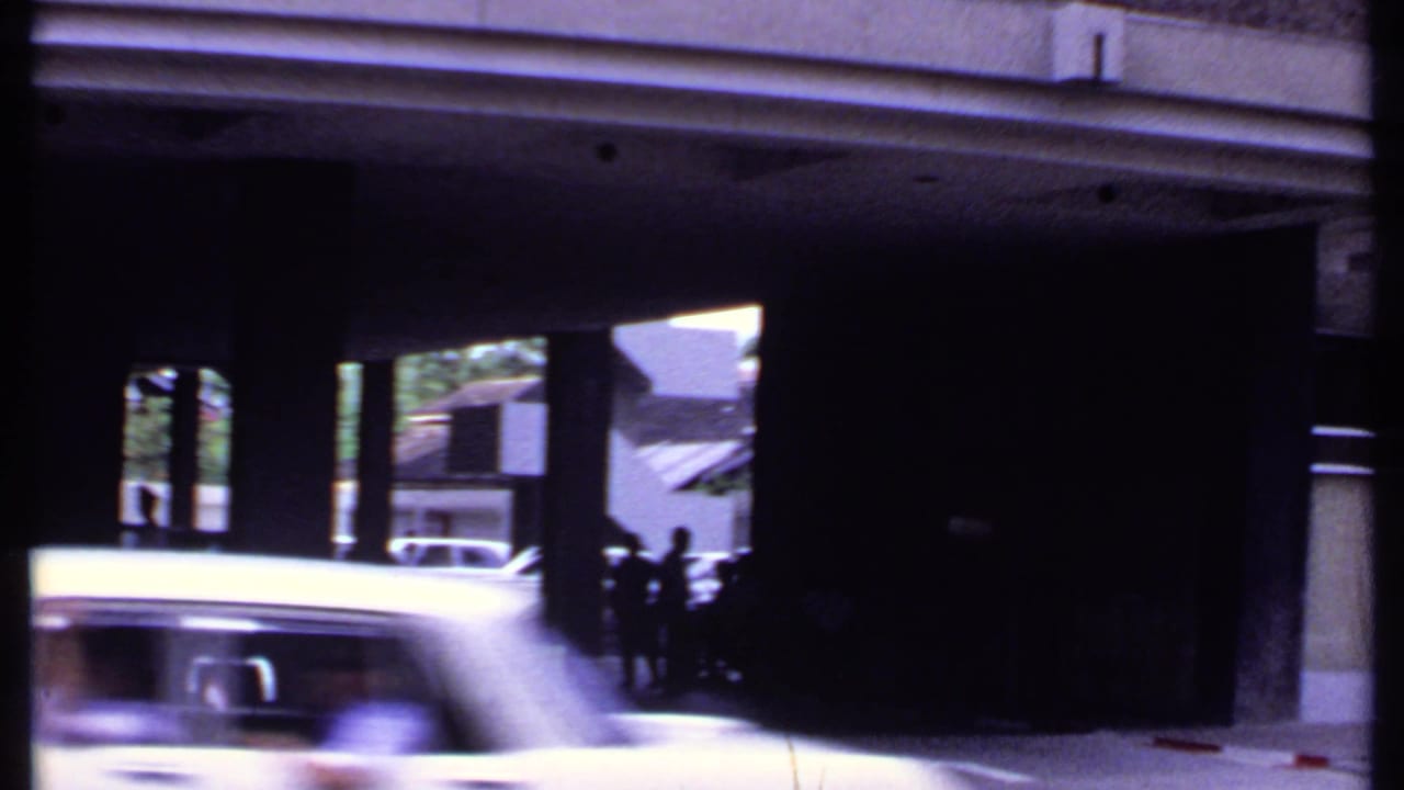1969 India 8mm street frame with a light-colored car crossing the foreground and people silhouetted near a modern building entrance.