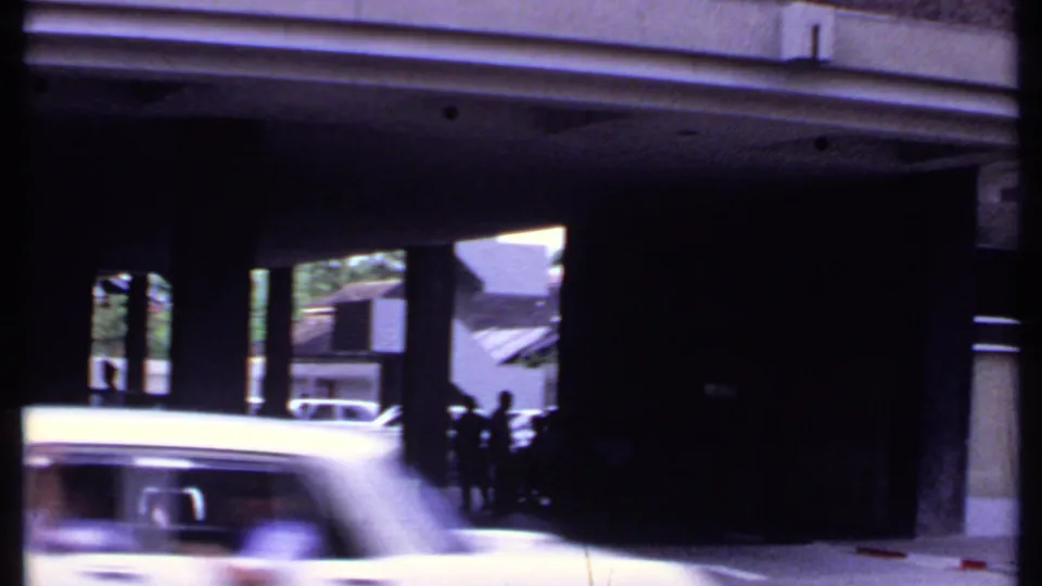 1969 India 8mm street frame with a light-colored car crossing the foreground and people silhouetted near a modern building entrance.