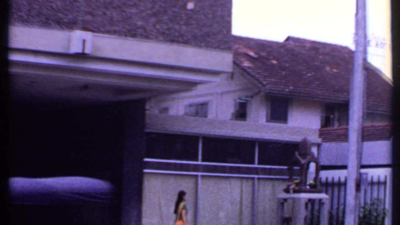 1969 India 8mm frame showing a tiled-roof residential-looking building, boundary wall, roadside pole, and a woman walking past.
