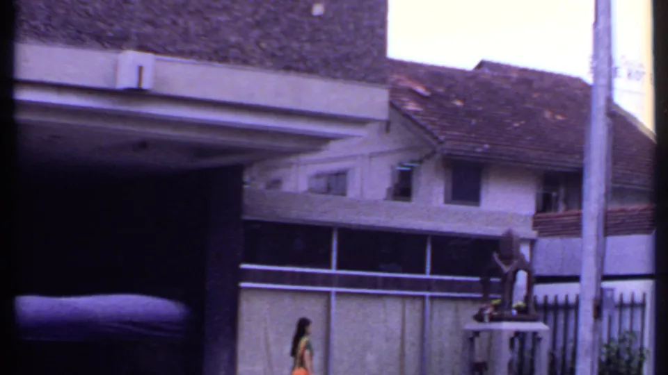 1969 India 8mm frame showing a tiled-roof residential-looking building, boundary wall, roadside pole, and a woman walking past.