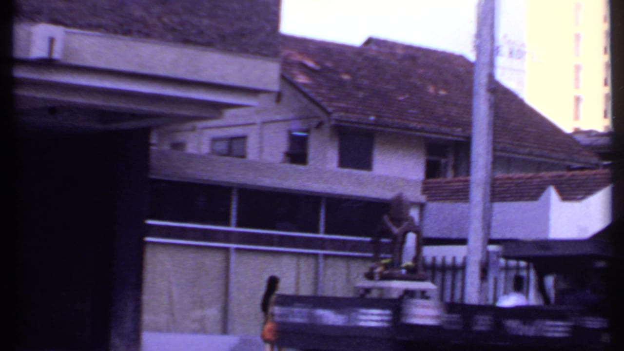 Street view with tiled-roof building, railings, poles, a statue-like feature, and a pedestrian in orange.