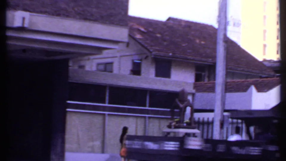 1969 India street frame showing tiled-roof buildings, roadside railings, vertical poles, and a pedestrian in orange near the curb.