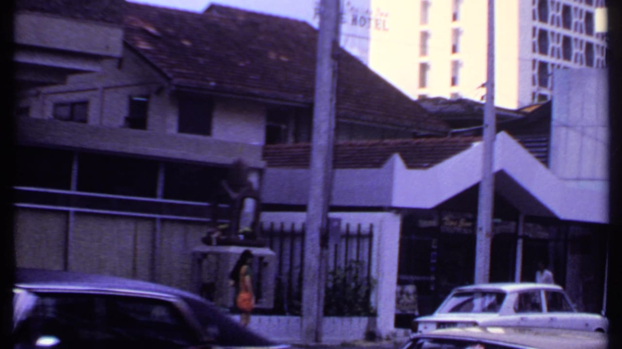 Street frontage with tiled roofs, hotel signage, a white sedan, a darker passing car, and a pedestrian.