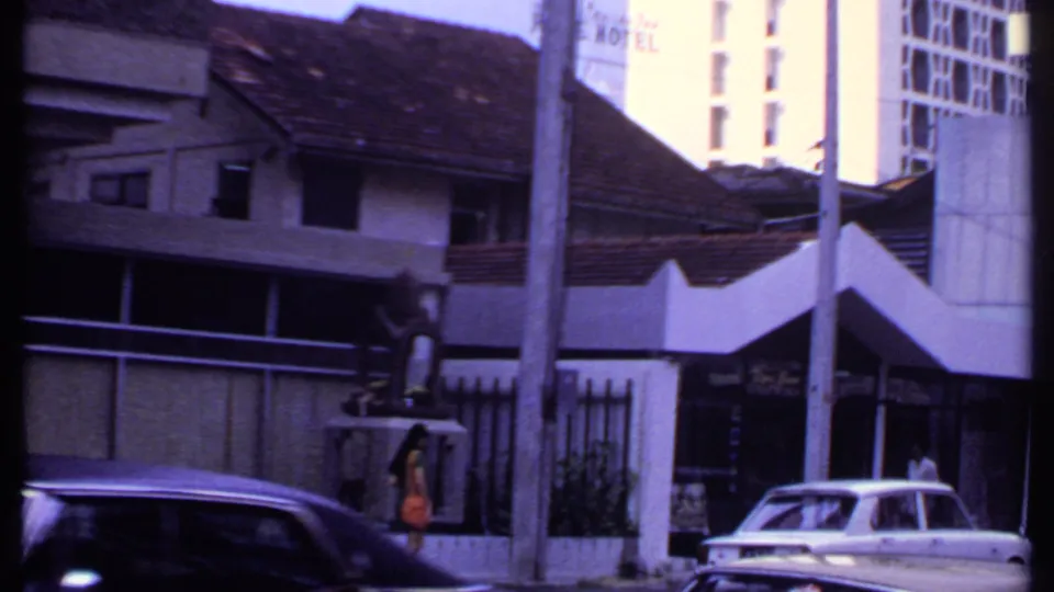 1969 India street scene with hotel signage, older tiled-roof buildings, a white sedan, a darker car, and pedestrians near the sidewalk.