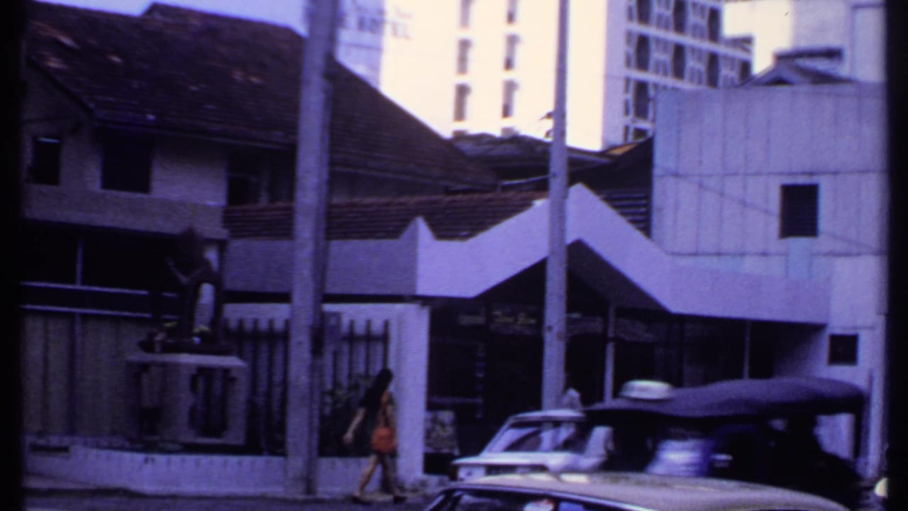 1969 India roadside frame with a pedestrian in orange, a white sedan, a dark vehicle, tiled roofs, poles, and a visible hotel sign.