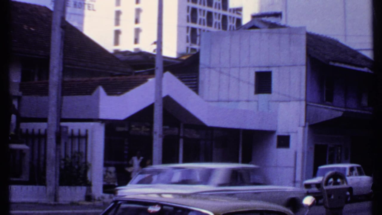 1969 India street frame with a white station wagon or sedan, older tiled roofs, a concrete facade, poles, hotel signage, and another car.