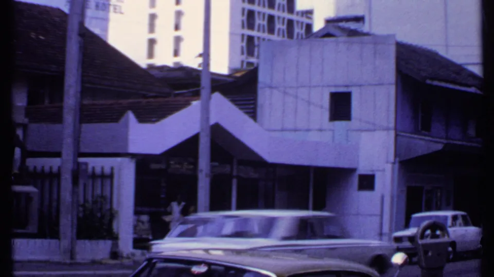 1969 India street frame with a white station wagon or sedan, older tiled roofs, a concrete facade, poles, hotel signage, and another car.
