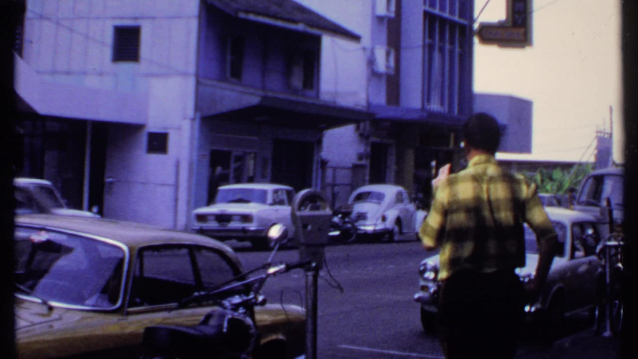 In 1969 India, a man in a plaid shirt walks away from camera near parked cars, a motorcycle, a parking meter, and street buildings.