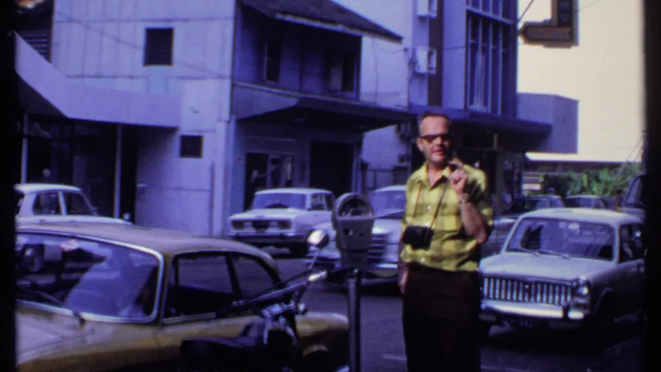 In 1969 India, a man wearing sunglasses and a yellow plaid shirt stands by a parking meter, with vintage cars and buildings behind him.