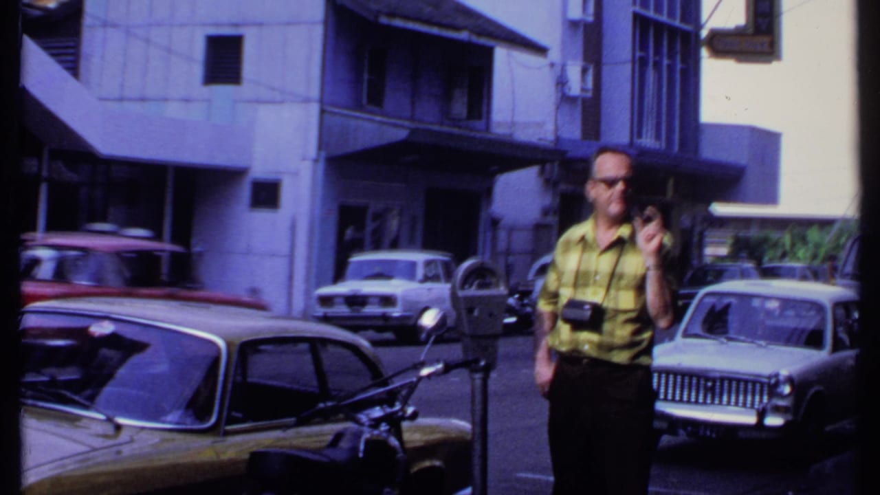 In 1969 India, the camera-carrying visitor stands near a parking meter while several vintage cars line the street around him.