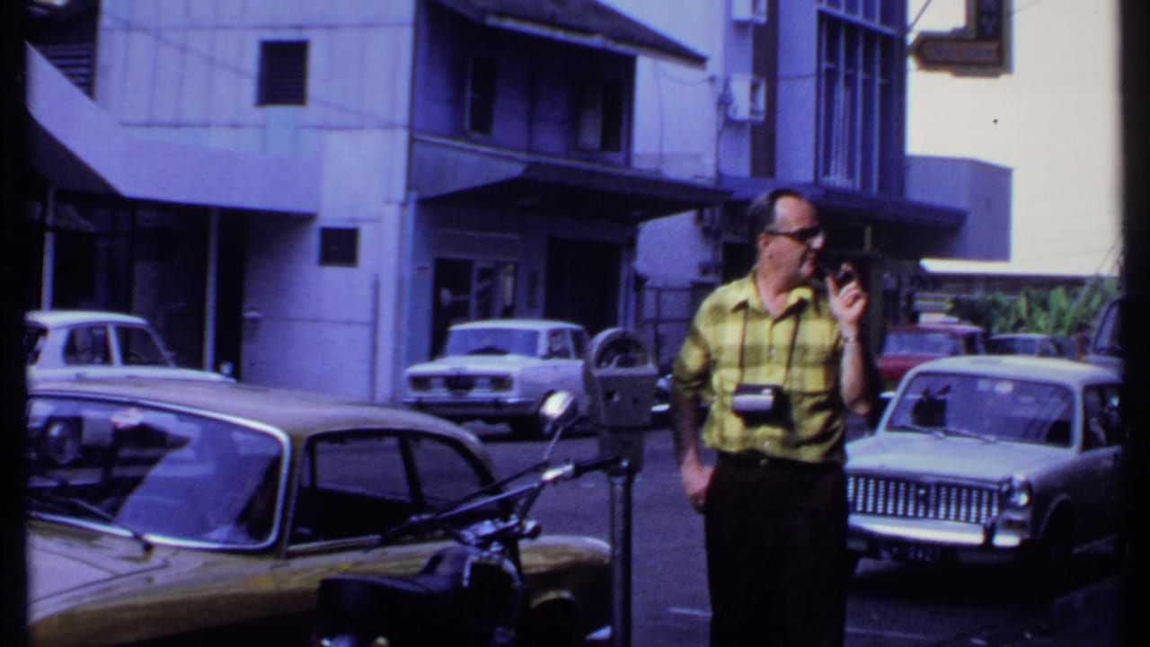 In 1969 India, a man in sunglasses and a yellow plaid shirt looks to the side near a parking meter, with vintage cars and buildings visible.