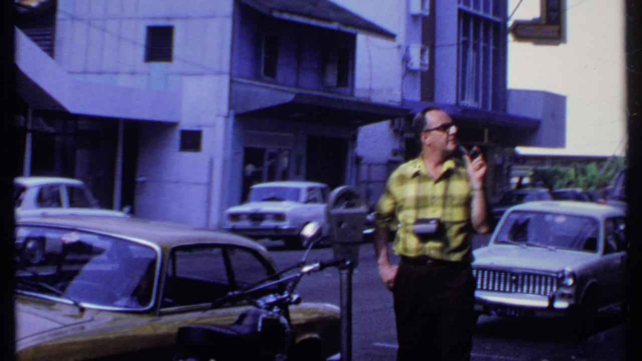 Man in yellow plaid shirt holding a camera on a street beside parked cars.