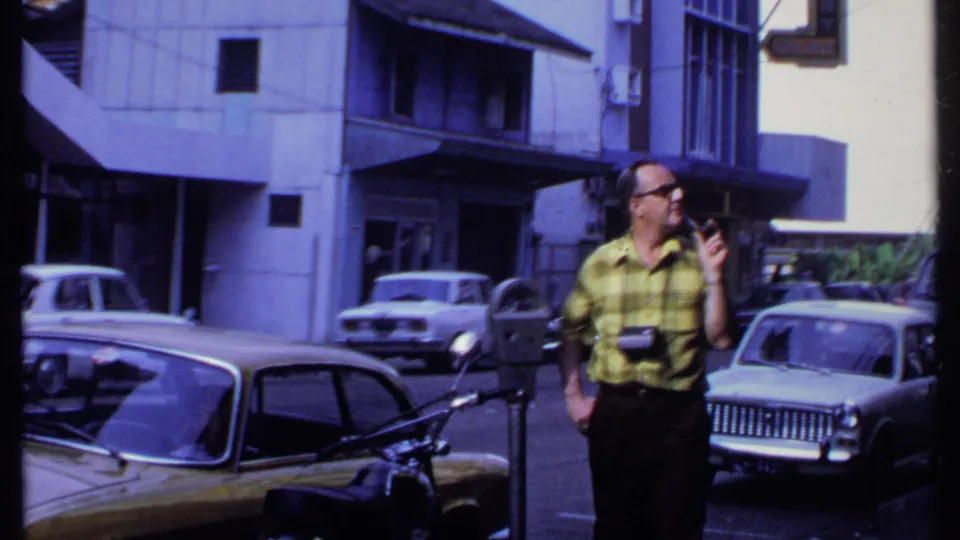 In 1969 India, a man with glasses and cameras stands on a sunlit street near parked cars, a motorcycle, and a parking meter.