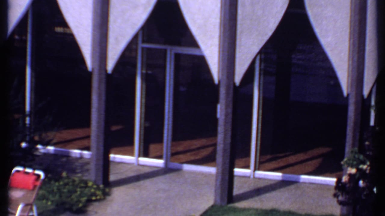 A modernist building facade in India shows triangular awnings, glass doors, planted areas, and outdoor chairs in bright sunlight.