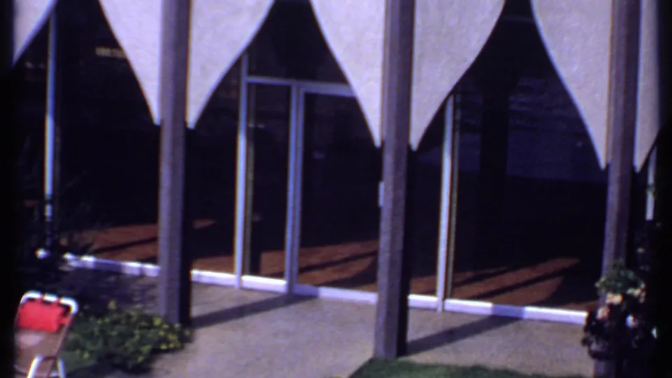 A modernist building facade in India shows triangular awnings, glass doors, planted areas, and outdoor chairs in bright sunlight.
