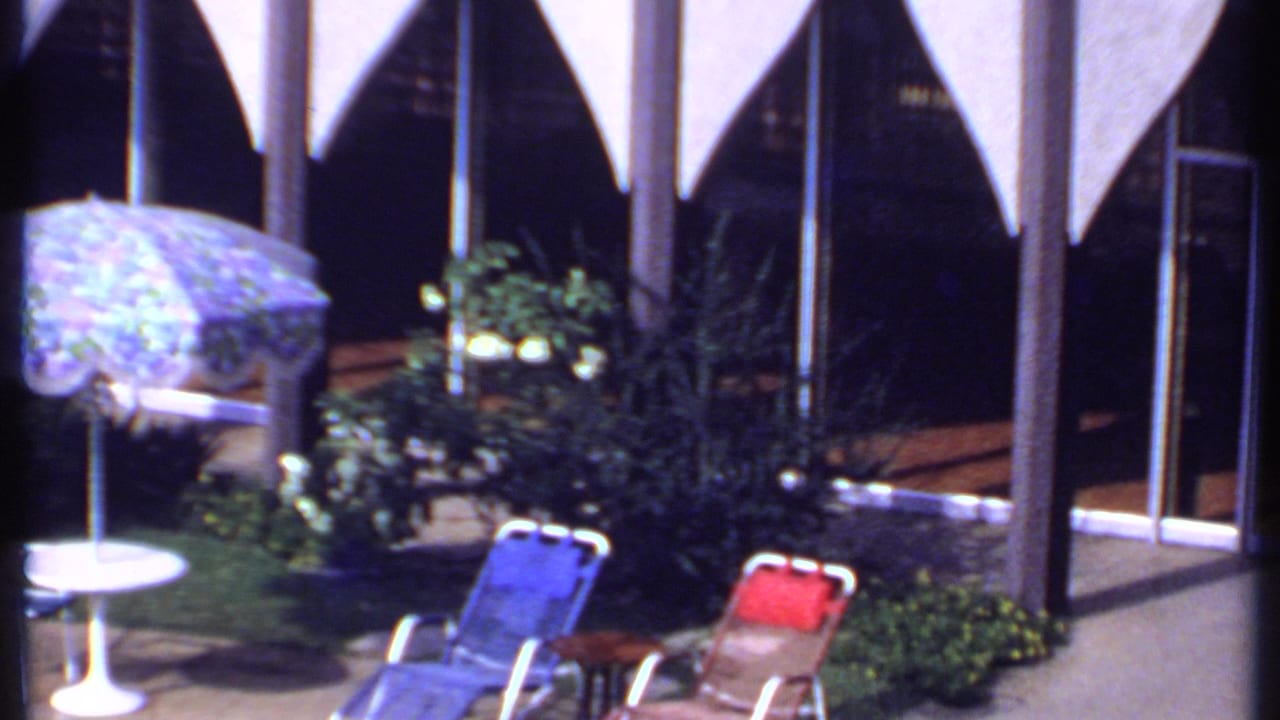 1969 India home-movie frame showing a poolside patio with a floral umbrella, white table, lounge chairs, plants, and arched architecture.
