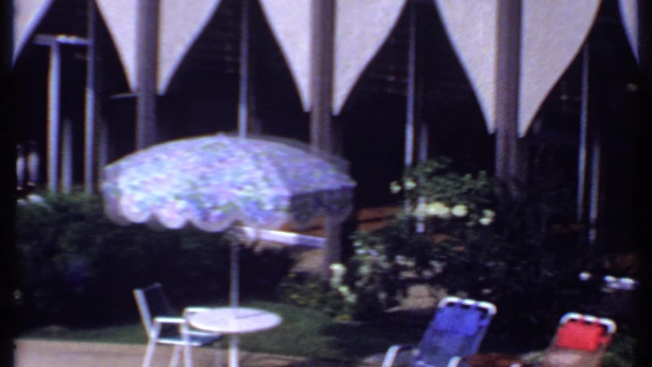 1969 India poolside patio view with a patterned umbrella, white table, chairs, shrubbery, and a modern arched building backdrop.