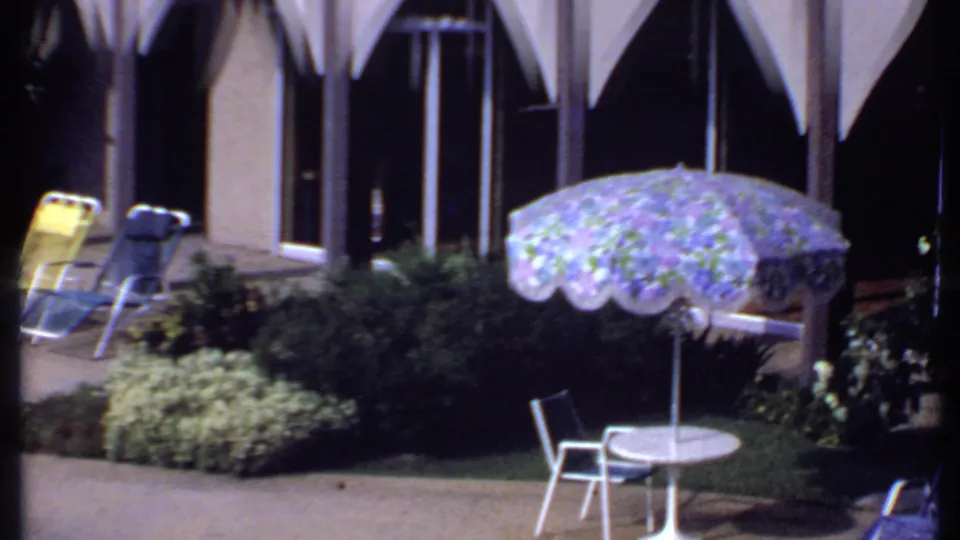 1969 India home-movie frame of lounge chairs and a floral umbrella near a walkway, landscaped shrubs, and glass doors.