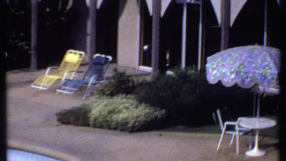 1969 India frame showing a landscaped poolside patio with colored lounge chairs, a floral umbrella, white table, and arched exterior.
