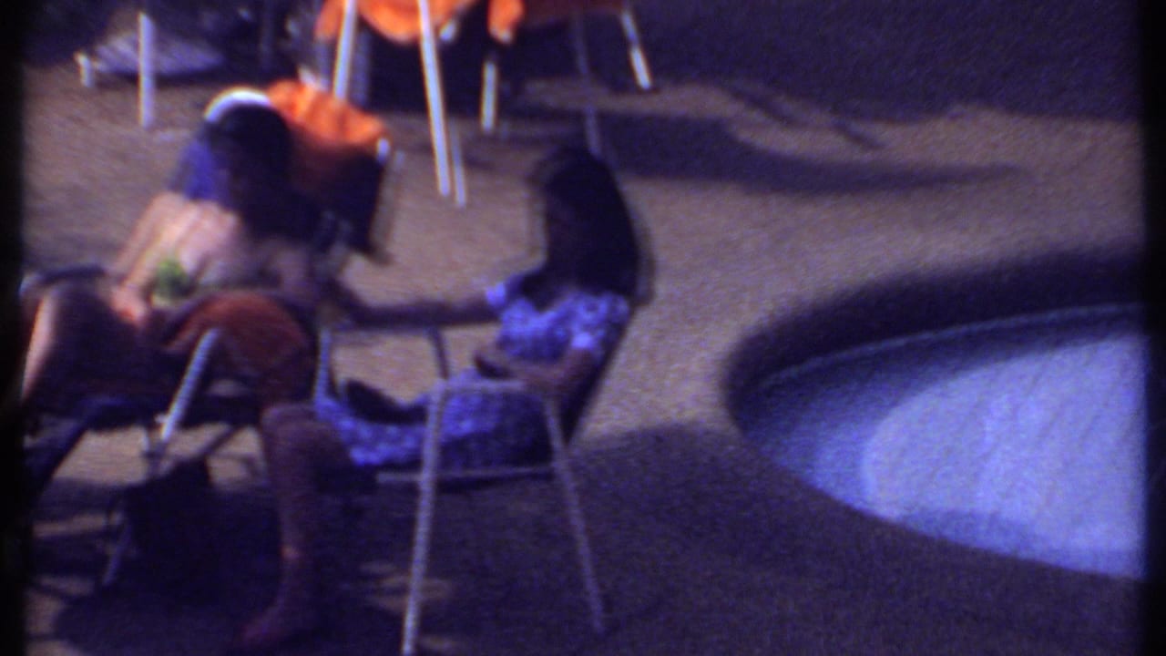 Adults seated in folding chairs beside a pool with orange towels and patio furniture visible.