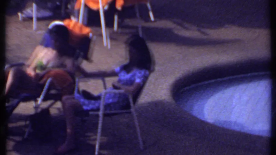 1969 India home-movie frame showing adults seated outdoors near a pool, with folding chairs, towels, and a casual patio setting.