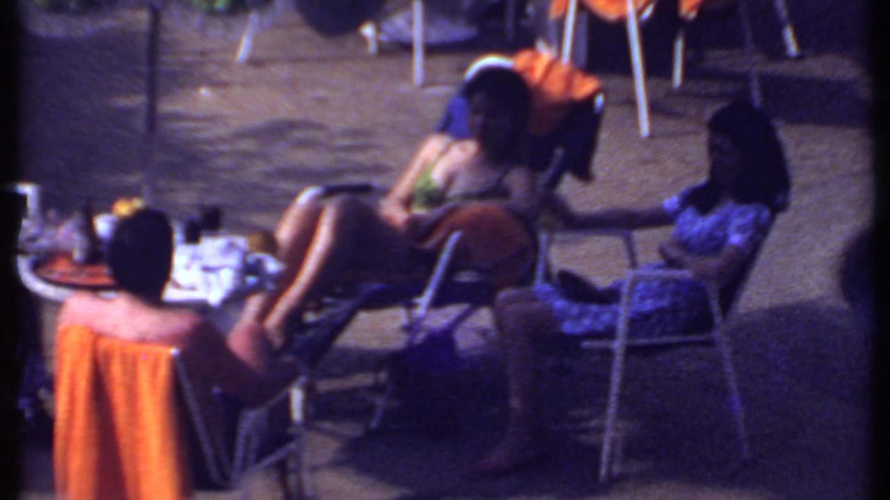 Small group seated around a round table outdoors with drinks, towels, and folding chairs.