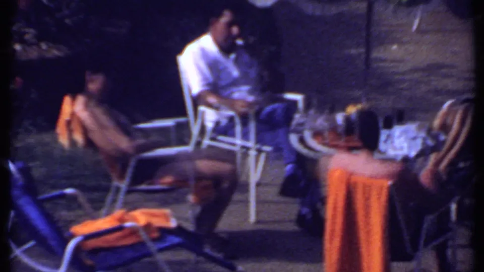 1969 India frame of adults conversing on patio chairs near a refreshment table, including a man in a white shirt and orange towels.