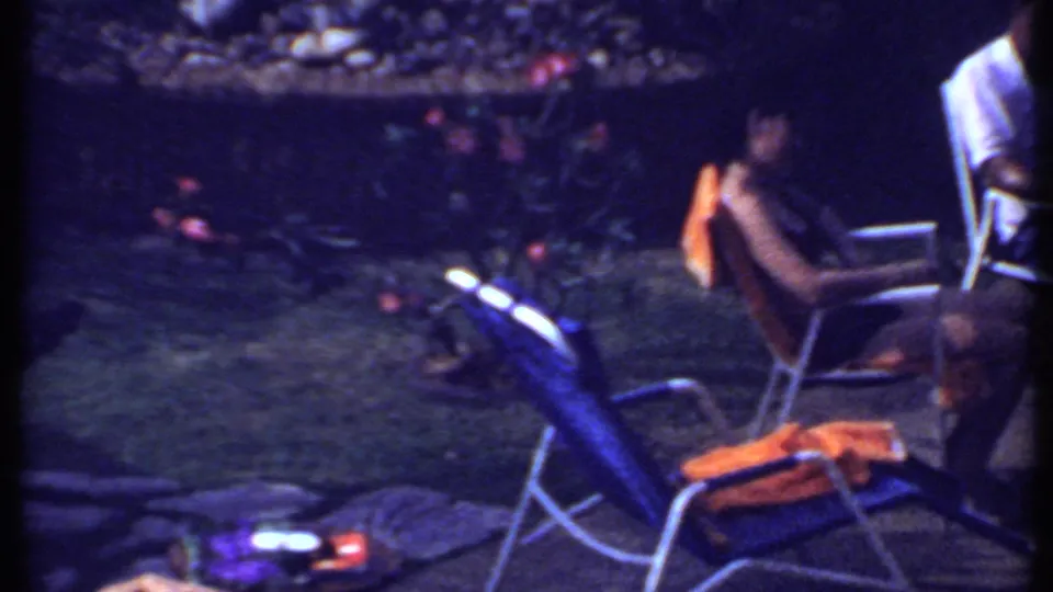 Poolside patio scene in India in 1969 with folding chairs, orange towels, garden planting, lawn, and adults partly visible.