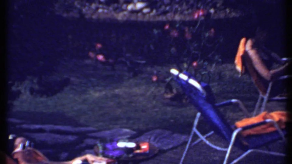 1969 India home-movie frame showing two women relaxing in an enclosed garden yard with lounge chairs and plants.