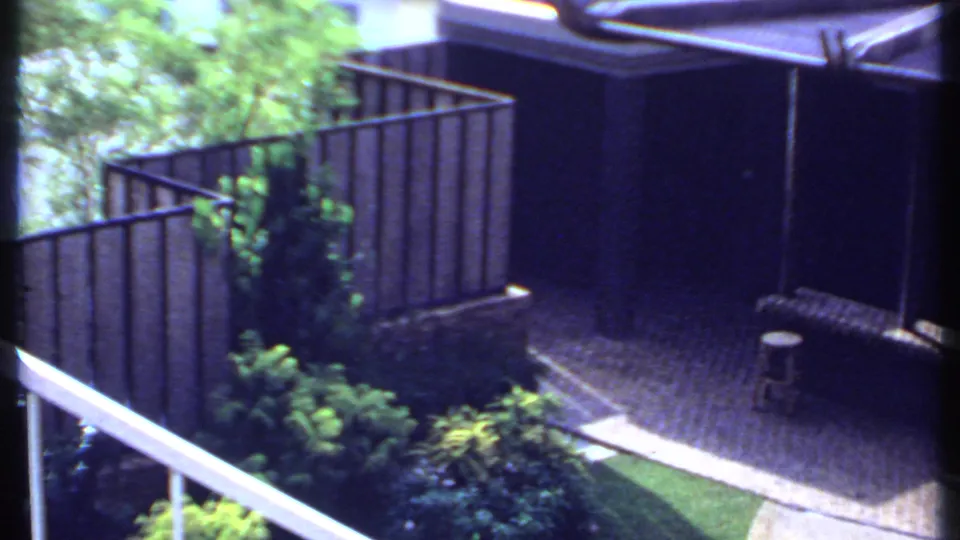 1969 India home-movie frame showing a fenced yard corner with paving, plants, railing, and outdoor seating.