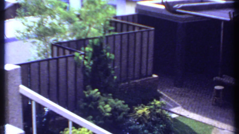 1969 India home-movie frame showing a modernist courtyard with greenery, metal railings, paving, and a shaded pergola.