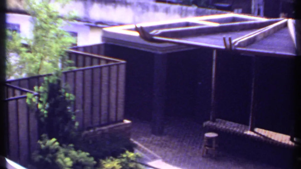 1969 India home-movie still showing a shaded pergola, paved courtyard, low railings, greenery, and a small stool.