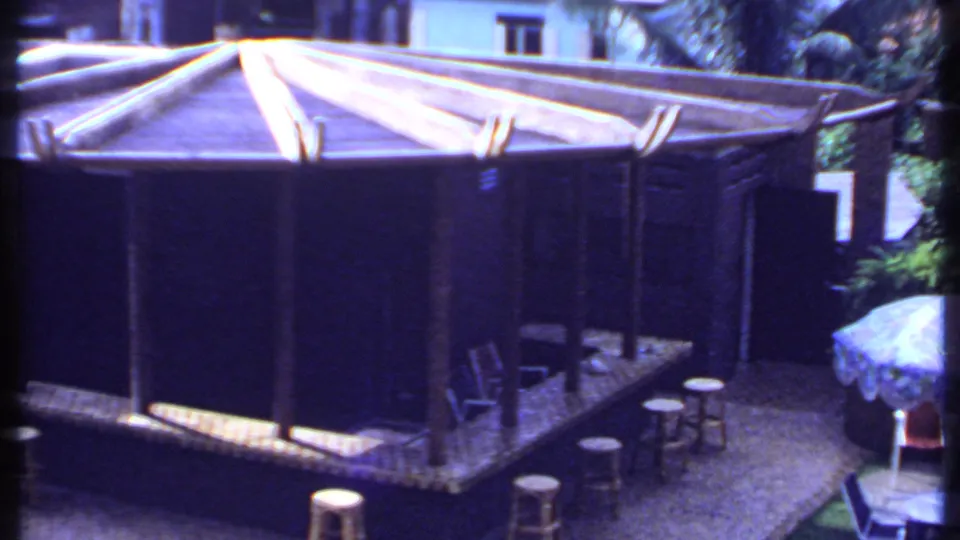 1969 India 8mm frame showing the bar structure beside garden greenery and a pale umbrella over a small outdoor table.