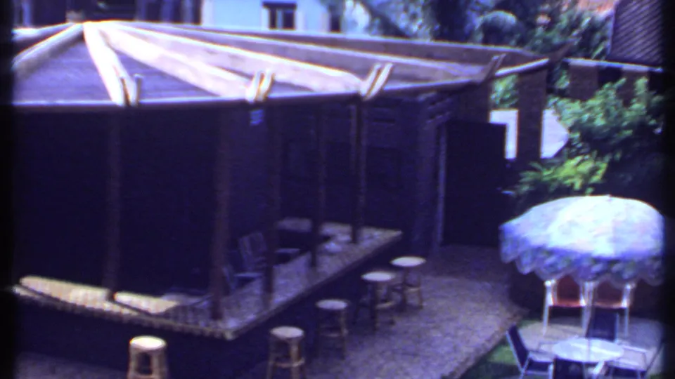 1969 India 8mm frame of the wooden garden bar with stools, surrounding plants, courtyard paving, and an umbrella-shaded table.